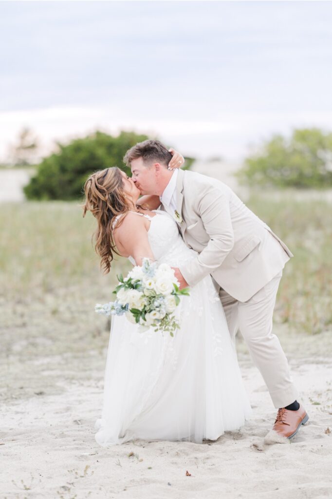 The groom dips the bride for a kiss during their beach wedding portraits while she holds her white and pale blue bouquet. The romantic pose is framed by sandy dunes and a soft evening sky.