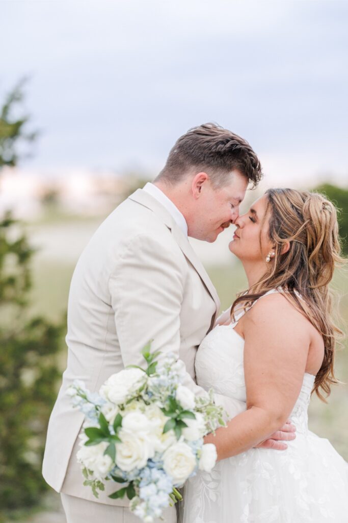 The bride and groom stand forehead to forehead, embracing during sunset portraits on the dunes. The bride’s bouquet of white and pale blue flowers adds a soft coastal touch to the intimate moment.