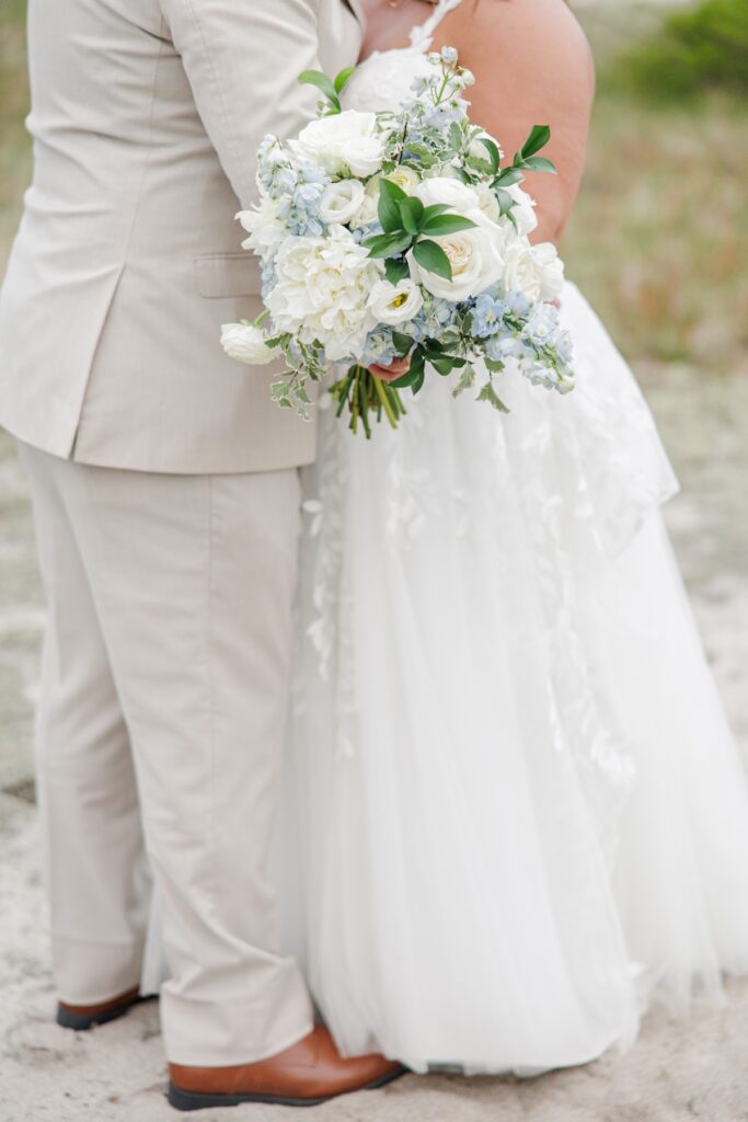 Close up of the bride’s bouquet featuring white roses, pale blue hydrangeas, ranunculus, delphinium, and fresh greenery against the couple’s wedding attire. The soft blue and white floral palette complements their Cape Cod beach wedding portraits.