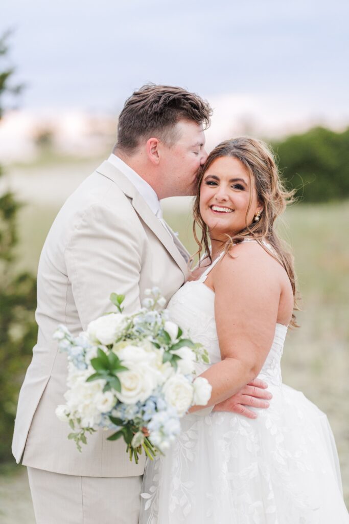 The groom kisses the bride’s forehead as she smiles toward the camera while holding her bouquet of white and pale blue flowers. The couple embraces among the dunes during golden hour portraits at Wychmere on Cape Cod.
