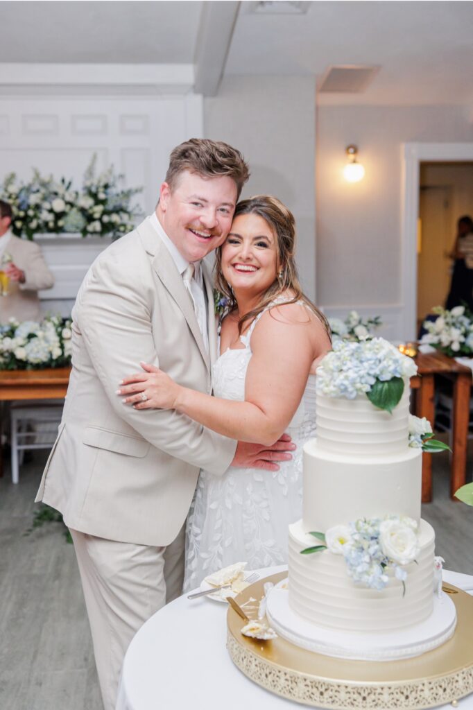 The bride and groom smile together beside their three tier white wedding cake decorated with white roses and pale blue hydrangeas. Floral arrangements throughout the Wychmere reception space continue the elegant coastal wedding design.