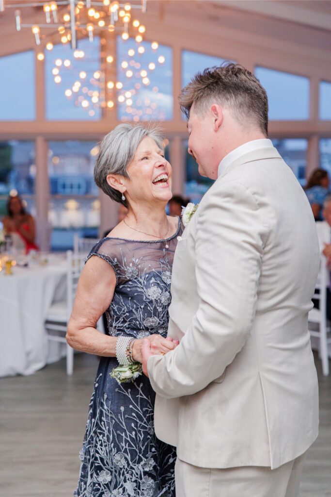The groom shares a joyful dance with a family member during the wedding reception at Wychmere. Soft chandelier lighting and waterfront windows create a warm atmosphere for the celebration.