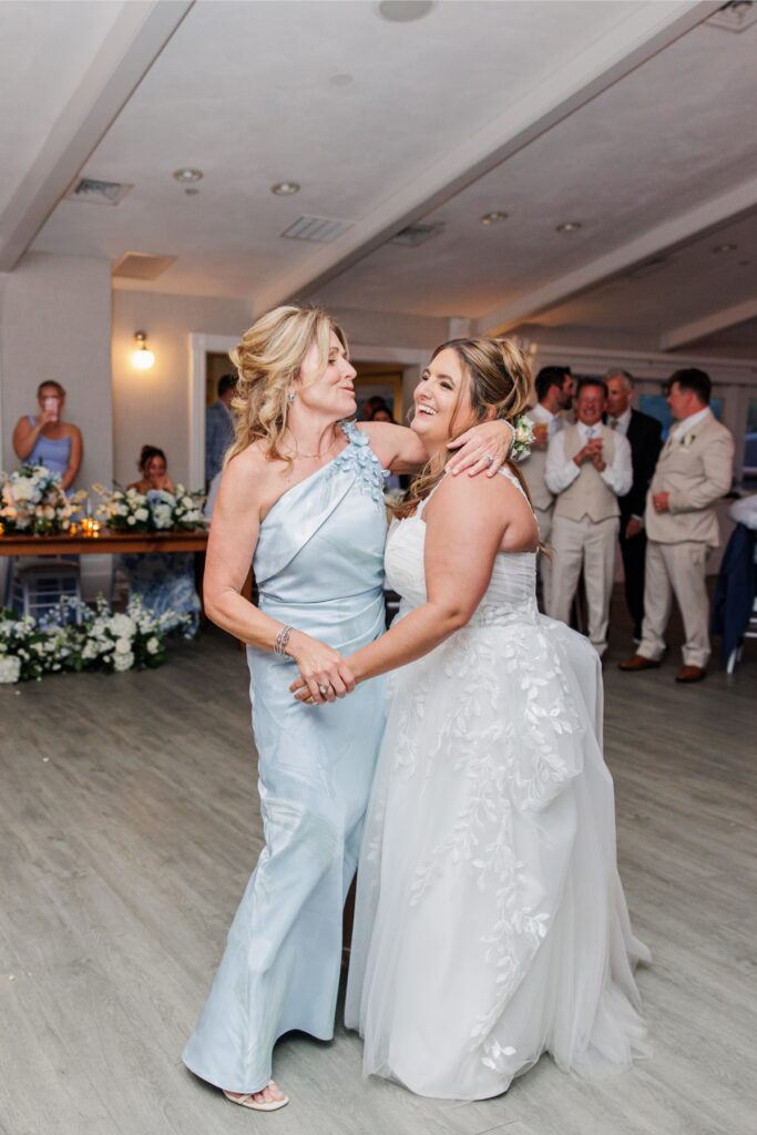 The bride shares a dance and laugh with a family member during the wedding reception while guests look on. White and pale blue floral arrangements decorate the reception space at Wychmere.