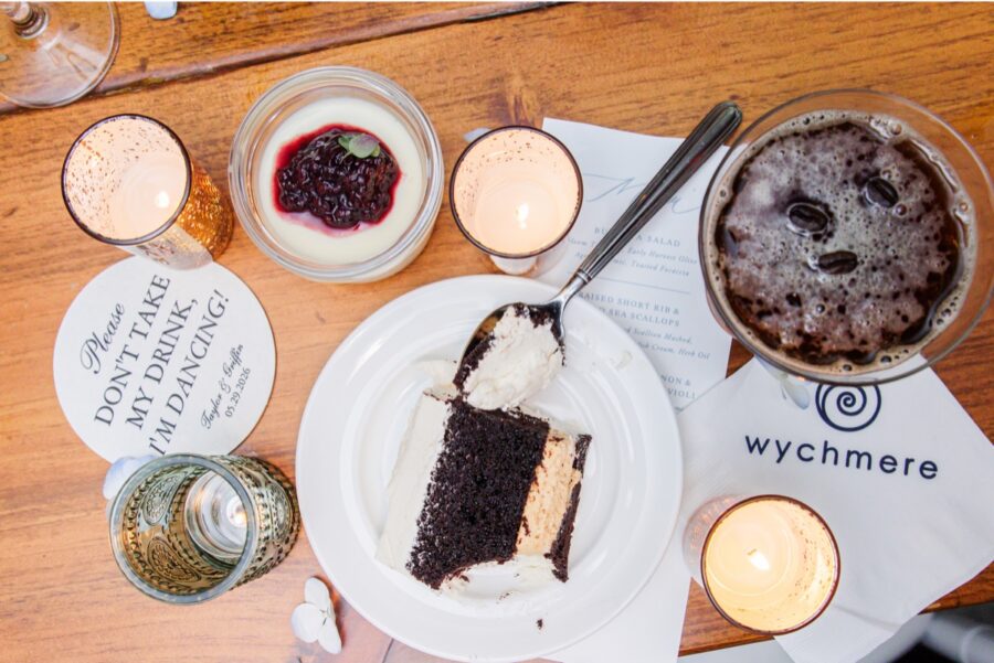 A slice of chocolate wedding cake topped with frosting sits beside dessert, candles, and a dark beer on a wooden reception table. A Wychmere cocktail napkin and personalized coaster add thoughtful details to the Cape Cod wedding celebration.