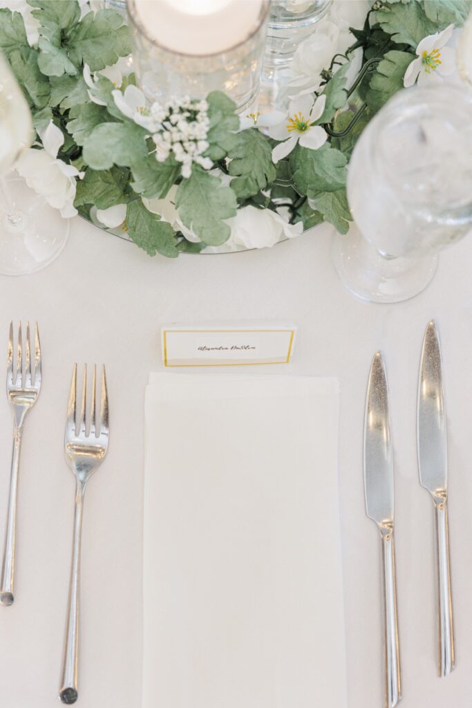 White wedding place setting with silver flatware, greenery, and a guest name card.