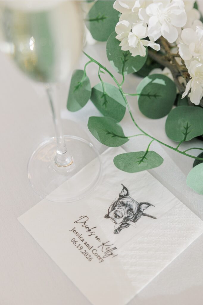 Wedding place setting with eucalyptus, white flowers, champagne, and a custom dog cocktail napkin.