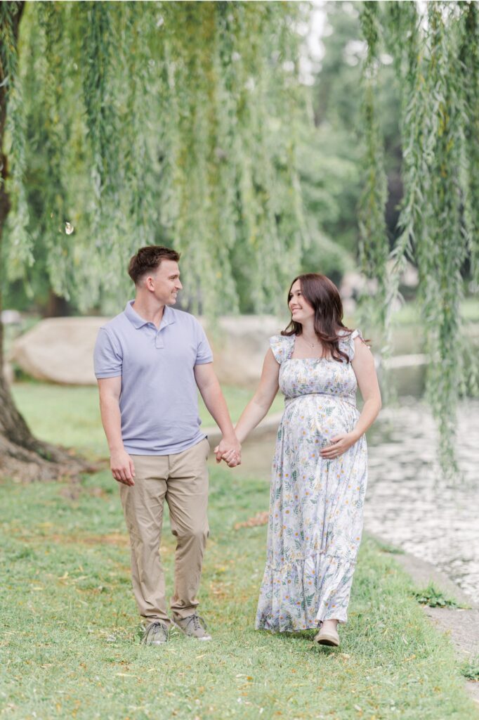 Couple holding hands walking by the lagoon at the Boston Public Garden during their maternity photos.