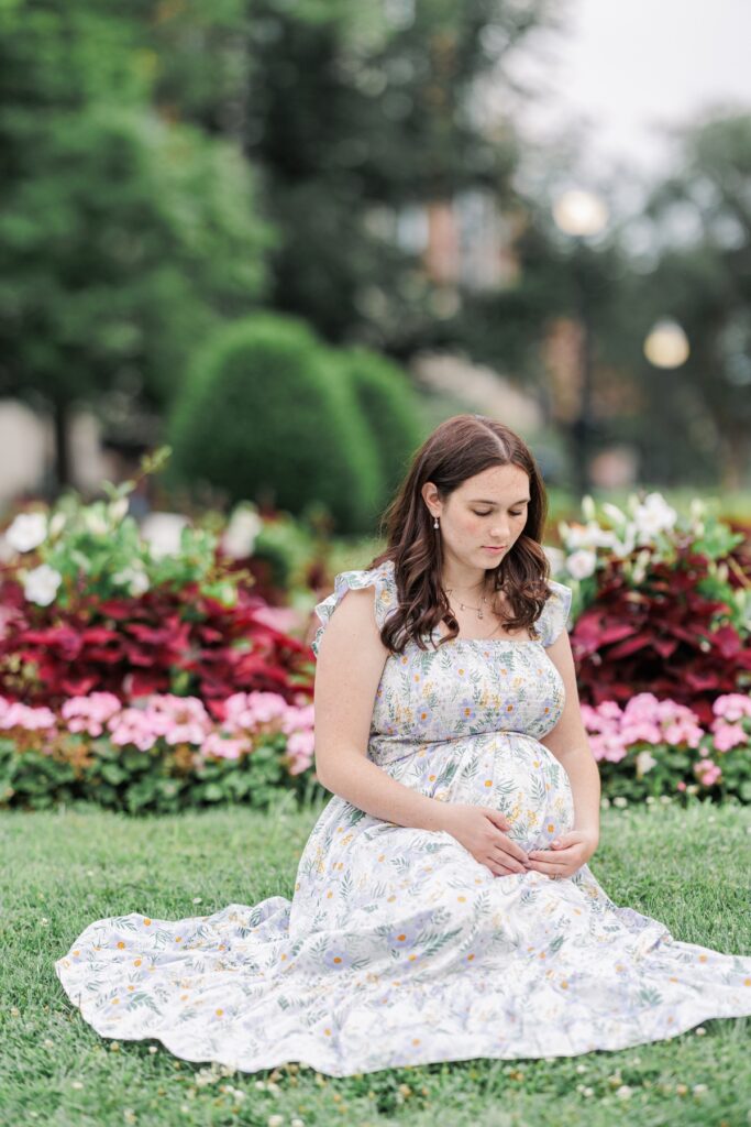A pregnant woman sitting in front of flowers looking down at her belly during her Boston Public Garden maternity photos