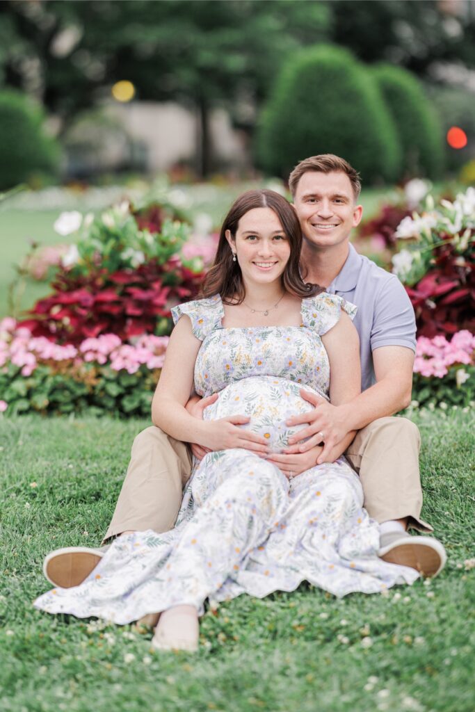 A couple sitting in front of flowers and smiling at the camera as they both hold the woman's pregnant belly during their Boston Public Garden maternity photo session.