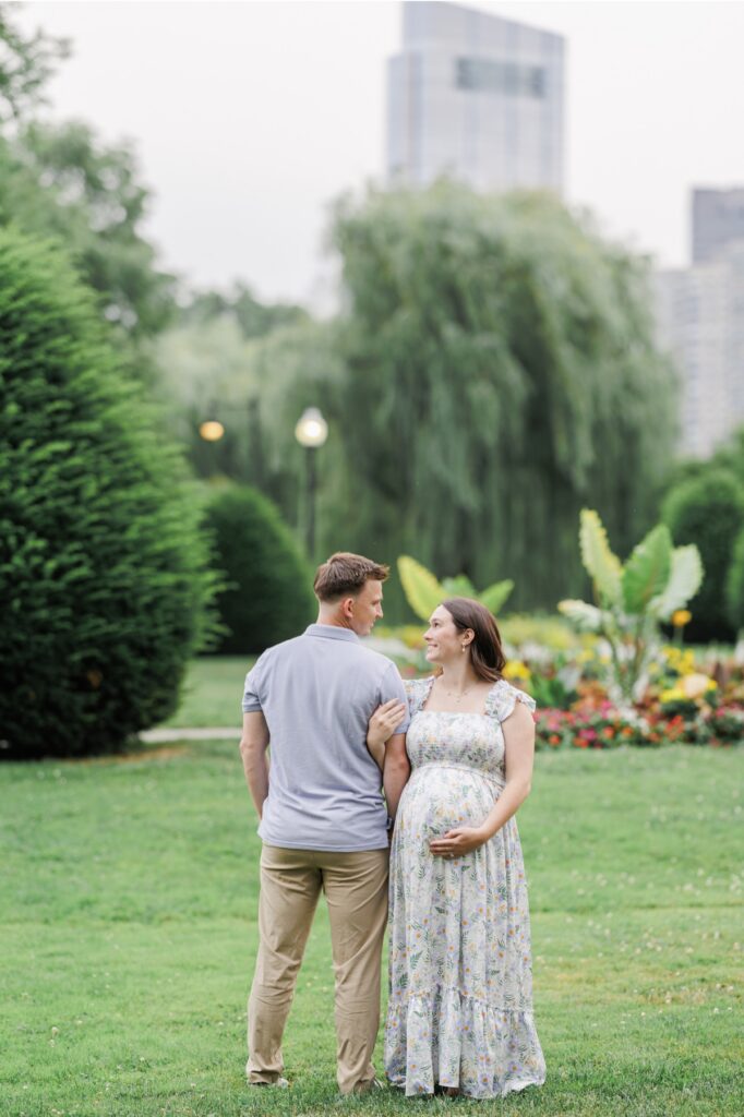 A pregnant woman facing the camera and turning her head to smile at her husband in the Boston Public Garden during their maternity photos. The man is facing away from the camera but smiling at his wife.