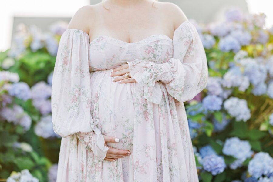 Close up of a pregnant woman holding her belly in front of hydrangea flowers.