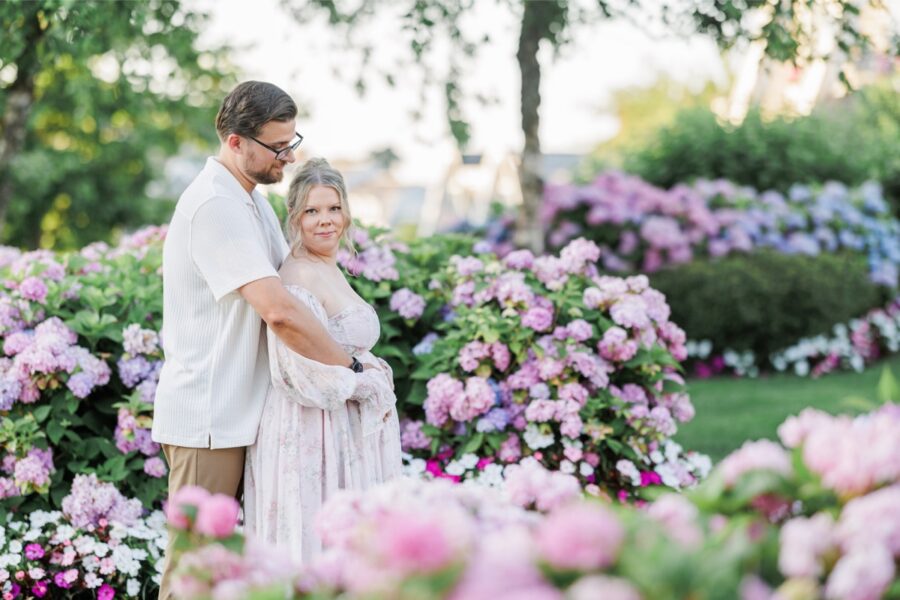 A couple hugging with the woman looking at the camera with a wall of hydrangea flowers behind them.