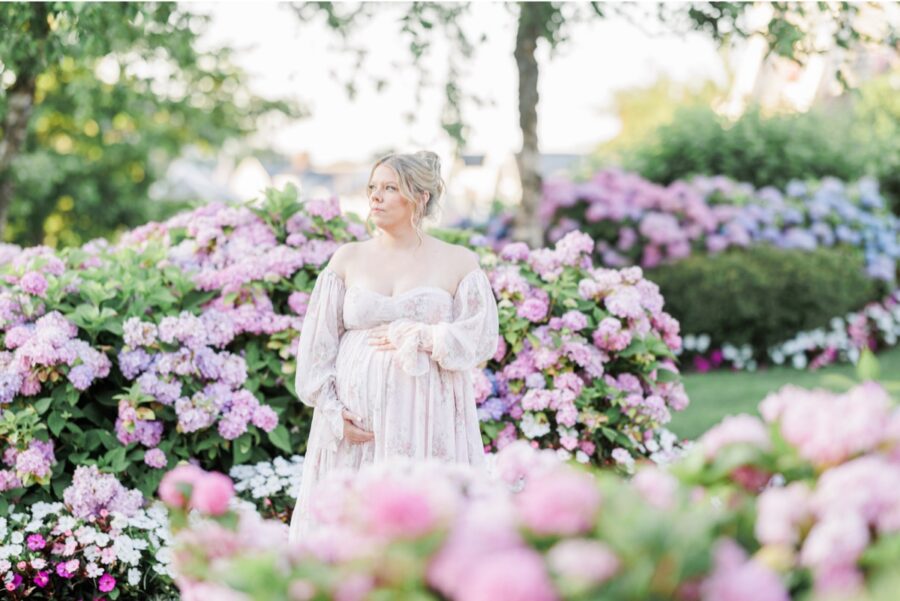 A pregnant woman holding her belly and looking into the distance while standing in front of hydrangea flowers.