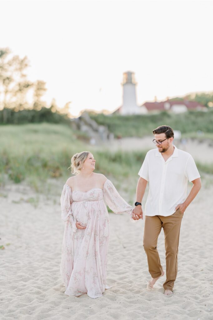 A couple holding hands and laughing while walking on the beach with Chatham Light in the background.