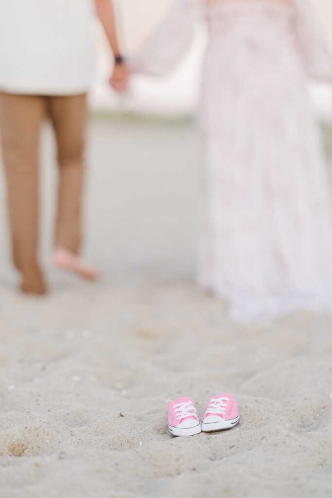 Pink baby shoes on the sand with a blurry couple walking away in the background