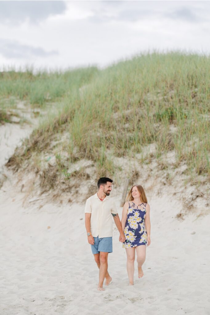 Amy and Joe walk hand in hand beside the beach grass during their Mayflower Beach engagement session.