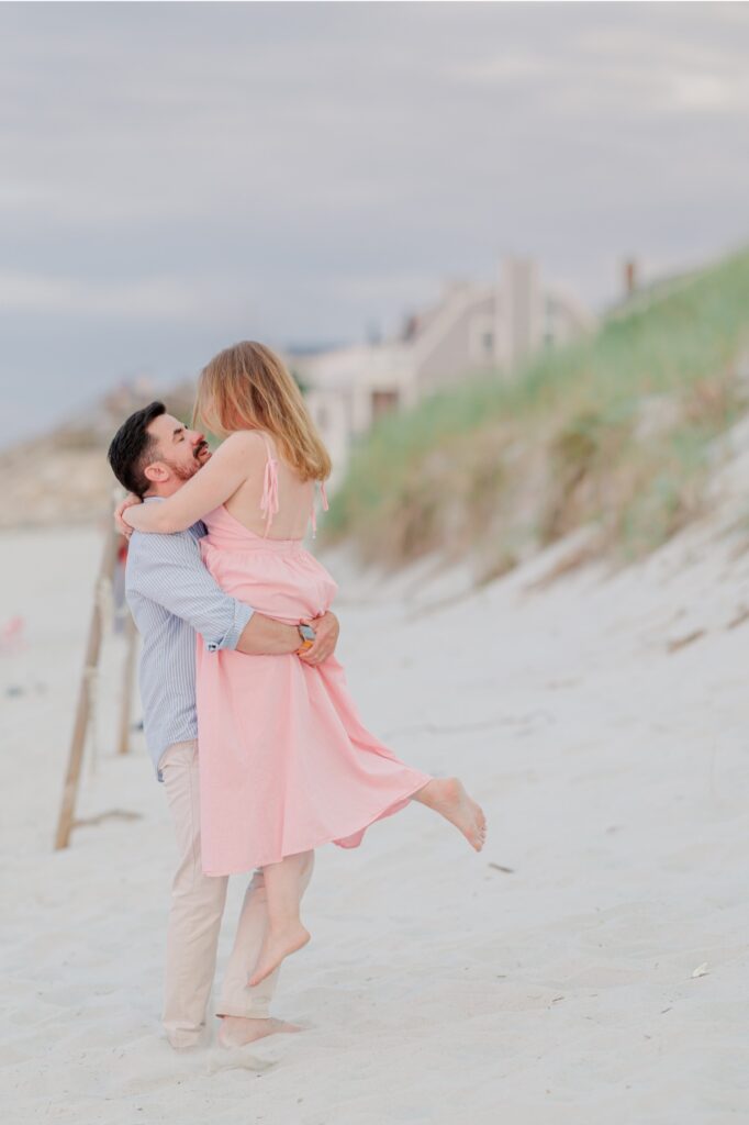 Joe lifts Amy beside the beach grass during their playful Mayflower Beach engagement session.