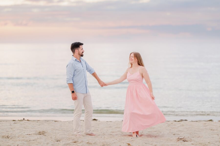 Amy and Joe hold hands on the sand beneath a soft pastel Cape Cod sky.