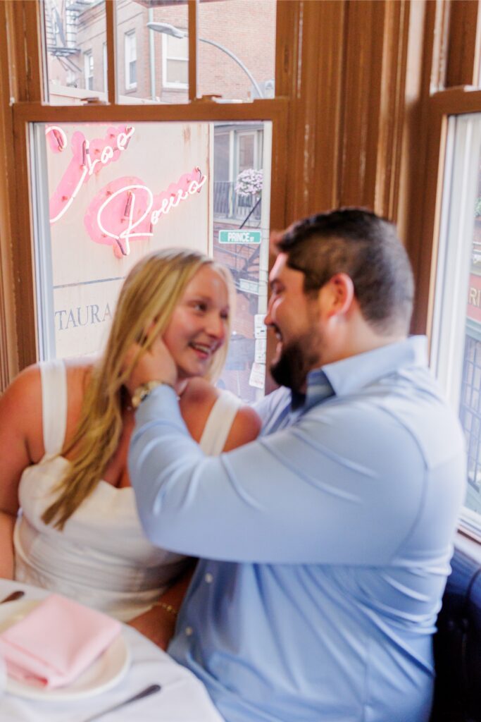 The engaged couple sitting by an upstairs window with the Ciao Roma sign visible in Boston’s North End