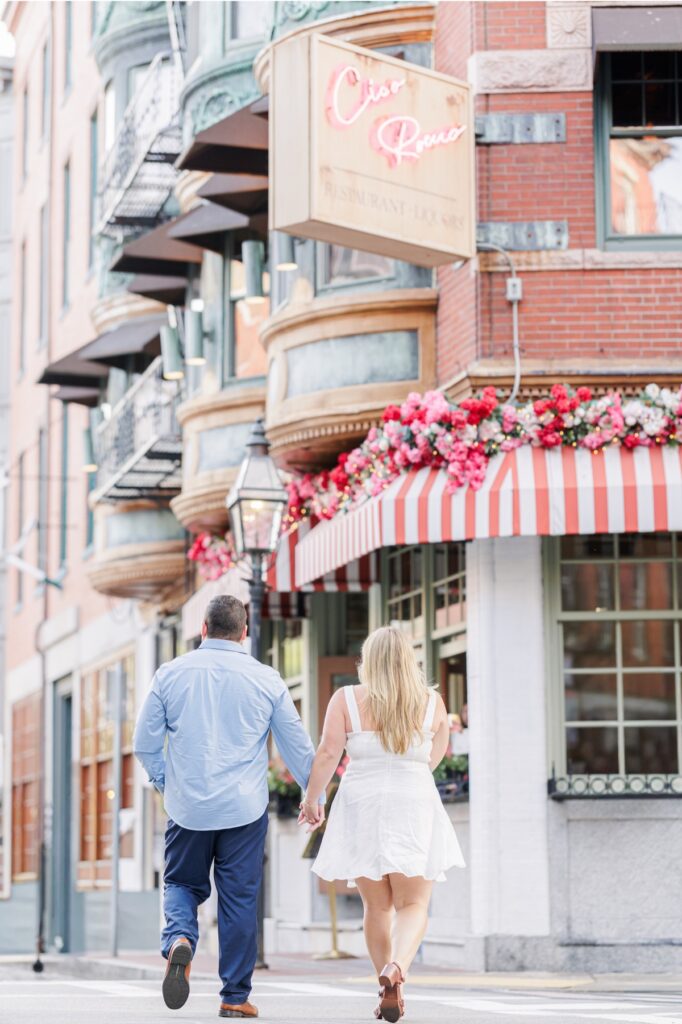 The engaged couple walking toward Ciao Roma beneath its striped awning in Boston’s North End
