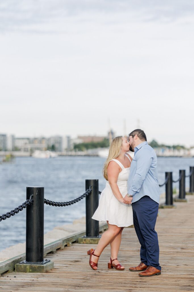 The engaged couple kissing along the Boston Harborwalk with the harbor behind them