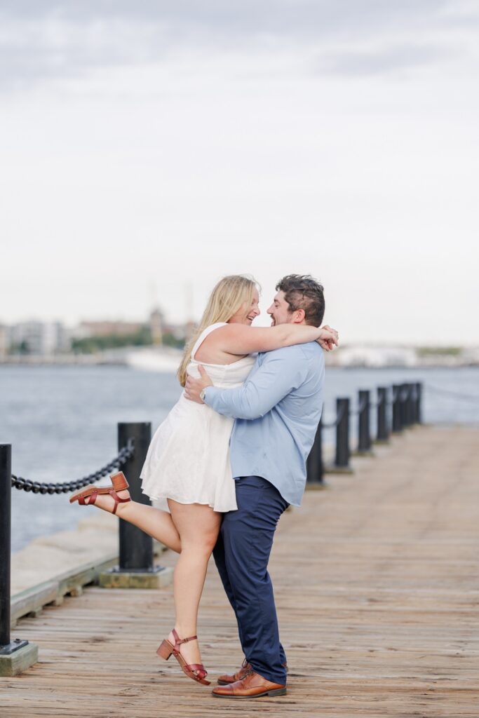 The engaged couple embracing playfully beside Boston Harbor on the Harborwalk