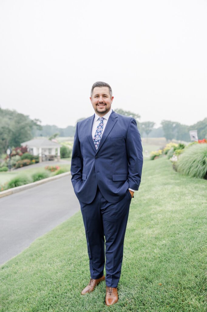 The groom in a navy suit and floral tie with the Salem Country Club wedding grounds behind him.