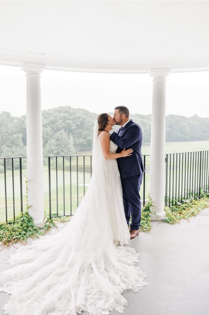 The bride and groom kissing between white porch columns at their rainy Salem Country Club wedding.