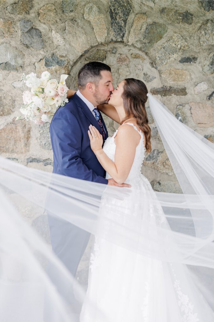 The bride and groom kissing against a stone wall with her veil flowing around them at their Salem Country Club wedding.