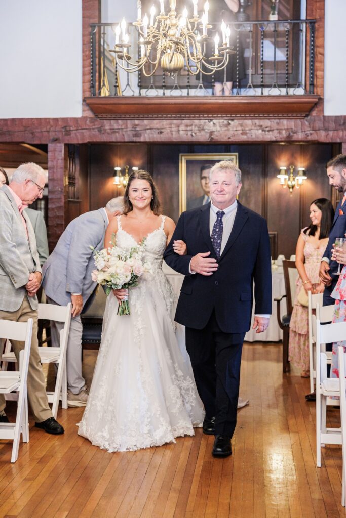 The bride walking down the aisle with her father during her indoor Salem Country Club wedding ceremony.