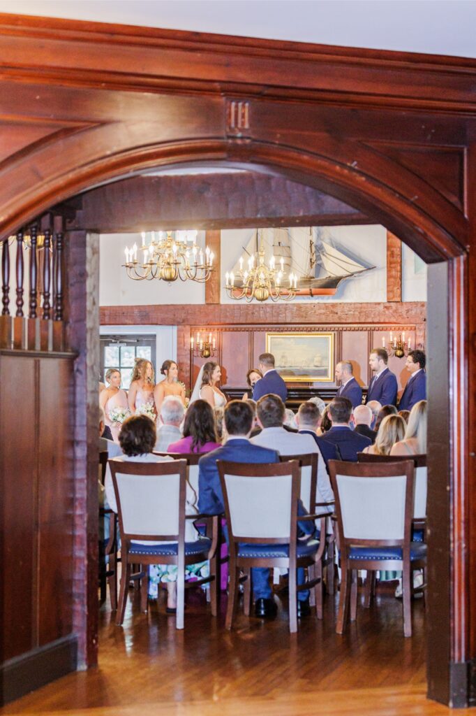 Guests watching the indoor Salem Country Club wedding ceremony beneath wood beams and brass chandeliers.