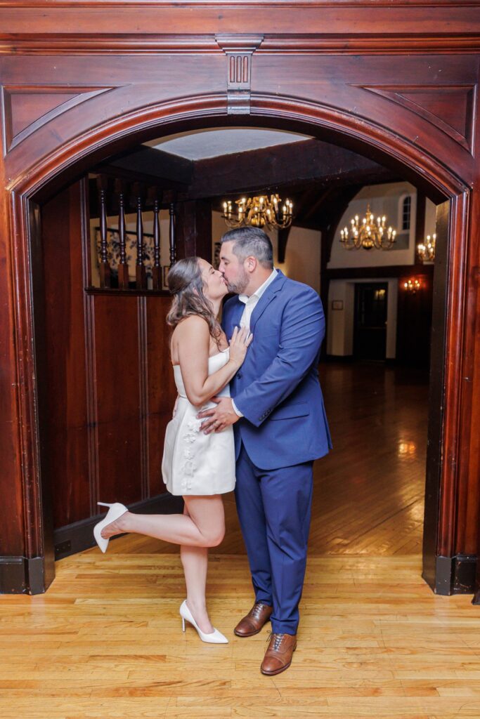 The bride in her short reception dress kissing the groom in a wooden doorway at their Salem Country Club wedding.