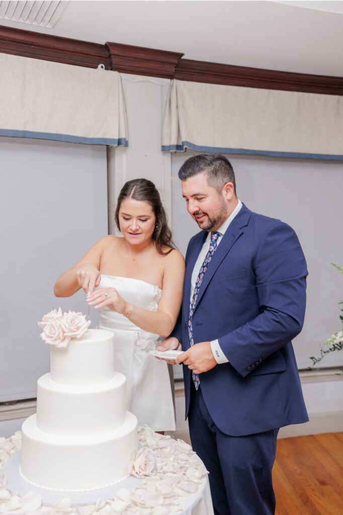 The bride cutting the white rose-topped cake as the groom holds a plate at their Salem Country Club wedding reception.