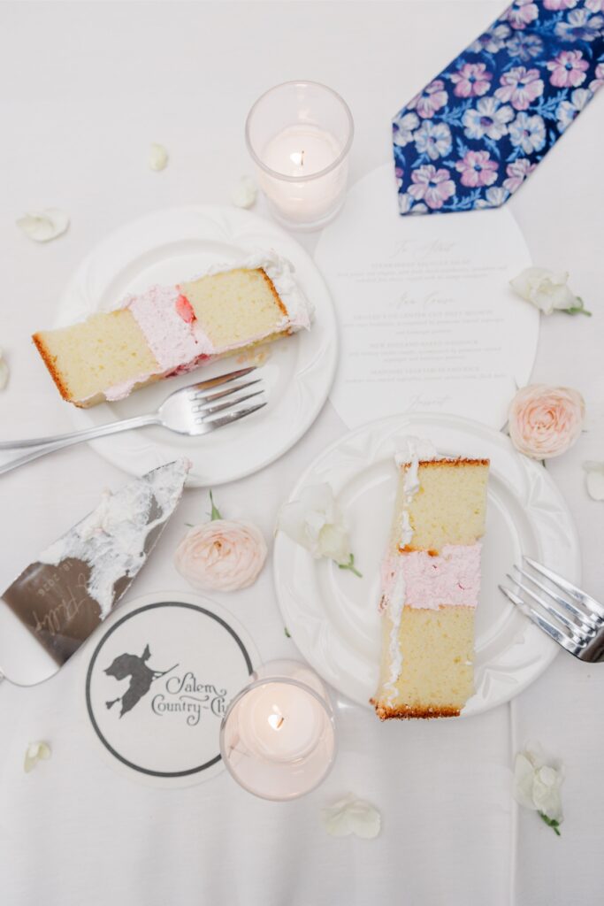 Two slices of wedding cake with pink filling on white plates among pale roses at the Salem Country Club wedding reception.