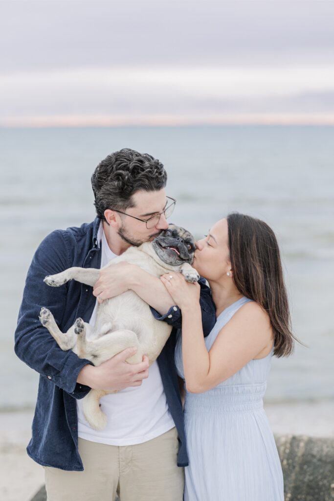 Couple kissing their pug while holding him close on the beach.