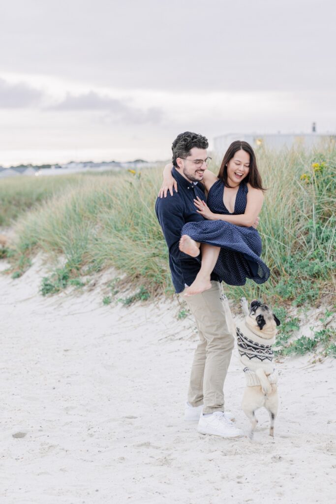 Man carrying his laughing fiancée beside beach grass with their pug nearby.